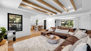 Un salon moderne avec un grand canapé d’angle, une table basse ronde et un tapis moelleux. On aperçoit à l’arrière-plan un coin repas avec une table et des chaises.