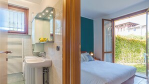 Ein Schlafzimmer mit einem großen Bett, ein Badezimmer mit Waschbecken und Spiegel sowie Blick auf Grünfläche.
