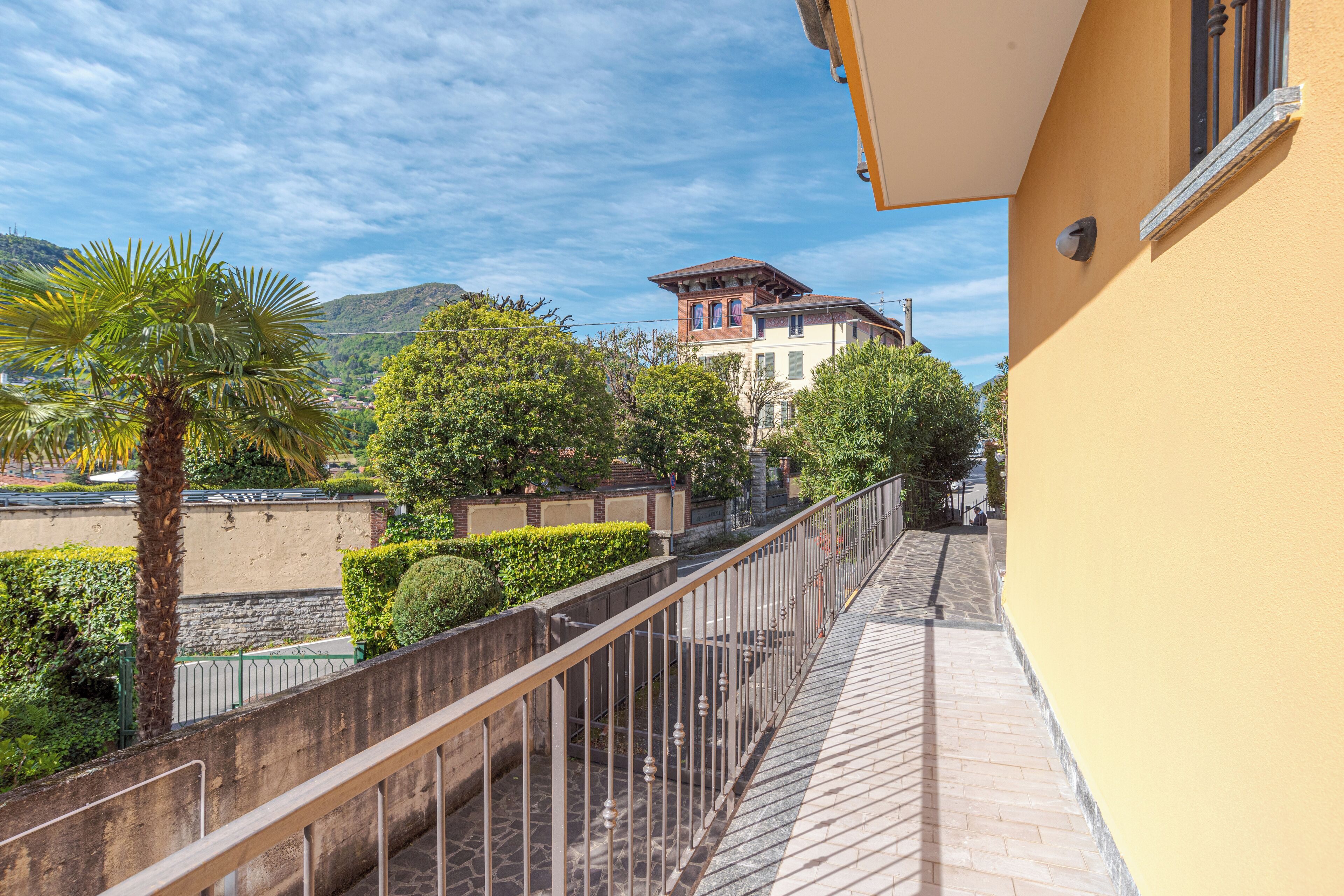 Un balcone con pavimento in pietra, ringhiera in metallo e vista su un edificio con tetto rosso e pareti bianche.