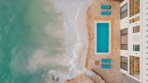 An aerial view of a beachfront property with a swimming pool, beach chairs, and a building with multiple windows.