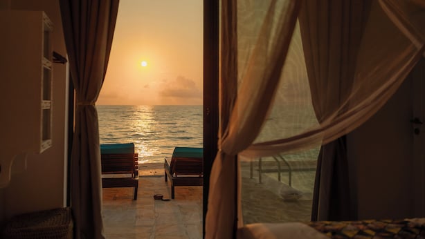 A room with a view of the sea at sunset, a bed, and two chairs.