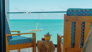 A balcony with two wooden chairs, one with a patterned cushion and the other with a solid color cushion. A wooden stool with a potted plant is in the center. The view includes the ocean and a clear sky.