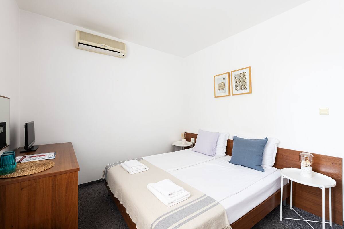 A hotel room with a bed, a desk, a chair, a small table, an air conditioner, and two framed pictures on the wall.