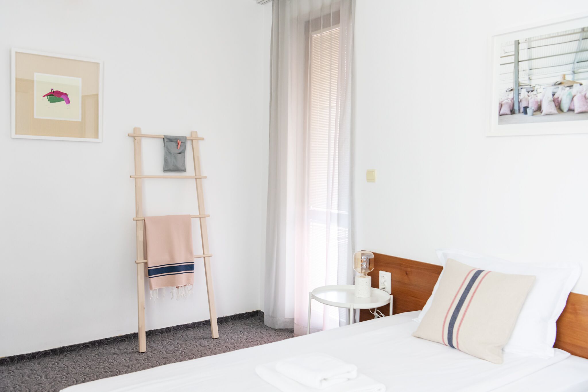 A hotel room with a bed, a bedside table with a lamp, a ladder, a towel, a framed picture on the wall, and a window with curtains.