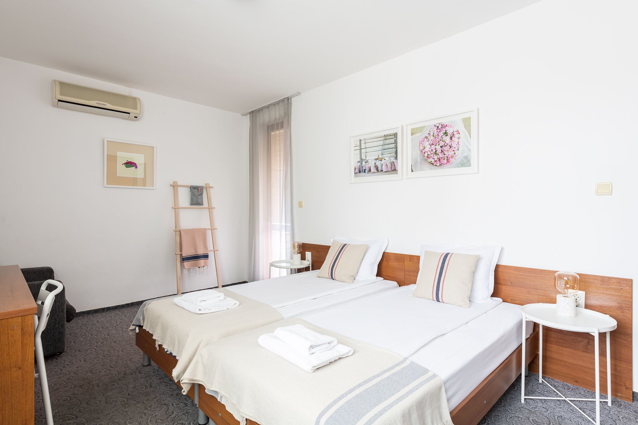 A hotel room with two beds, a desk, a chair, a small table, and a wall-mounted air conditioner.