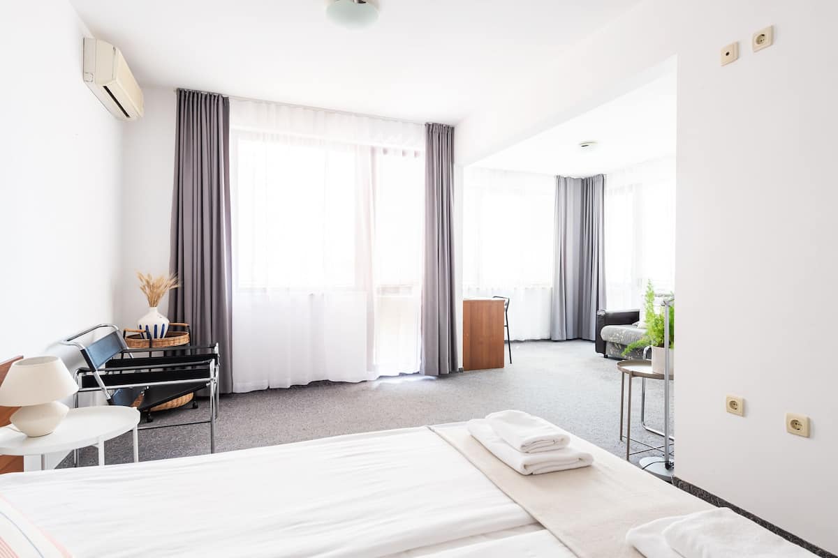 A hotel room with a bed, a desk, a chair, a small table with a plant, and a window with curtains.
