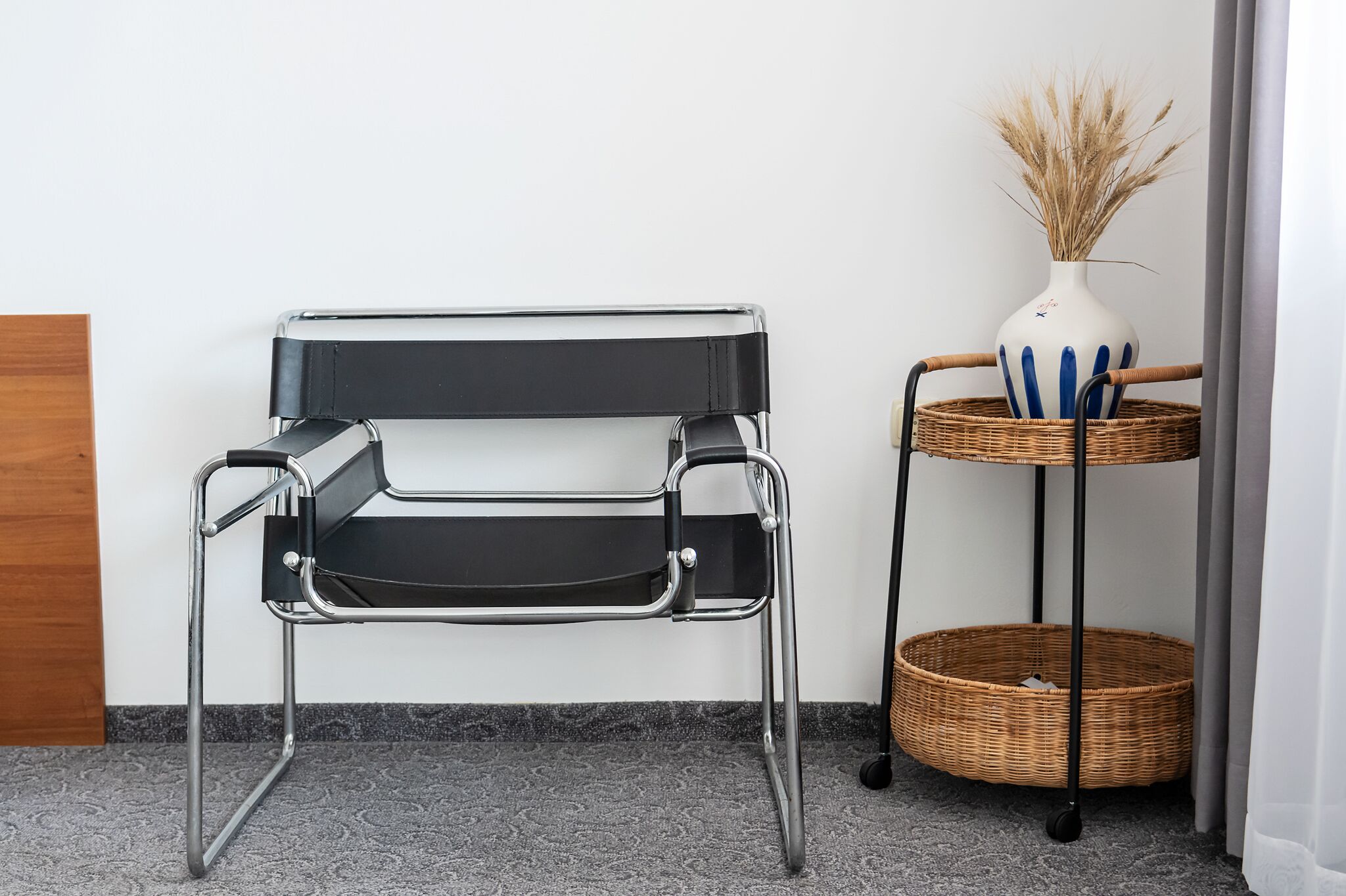 A black metal chair with a chrome frame, a wicker basket on a metal stand, and a white vase with dried plants.