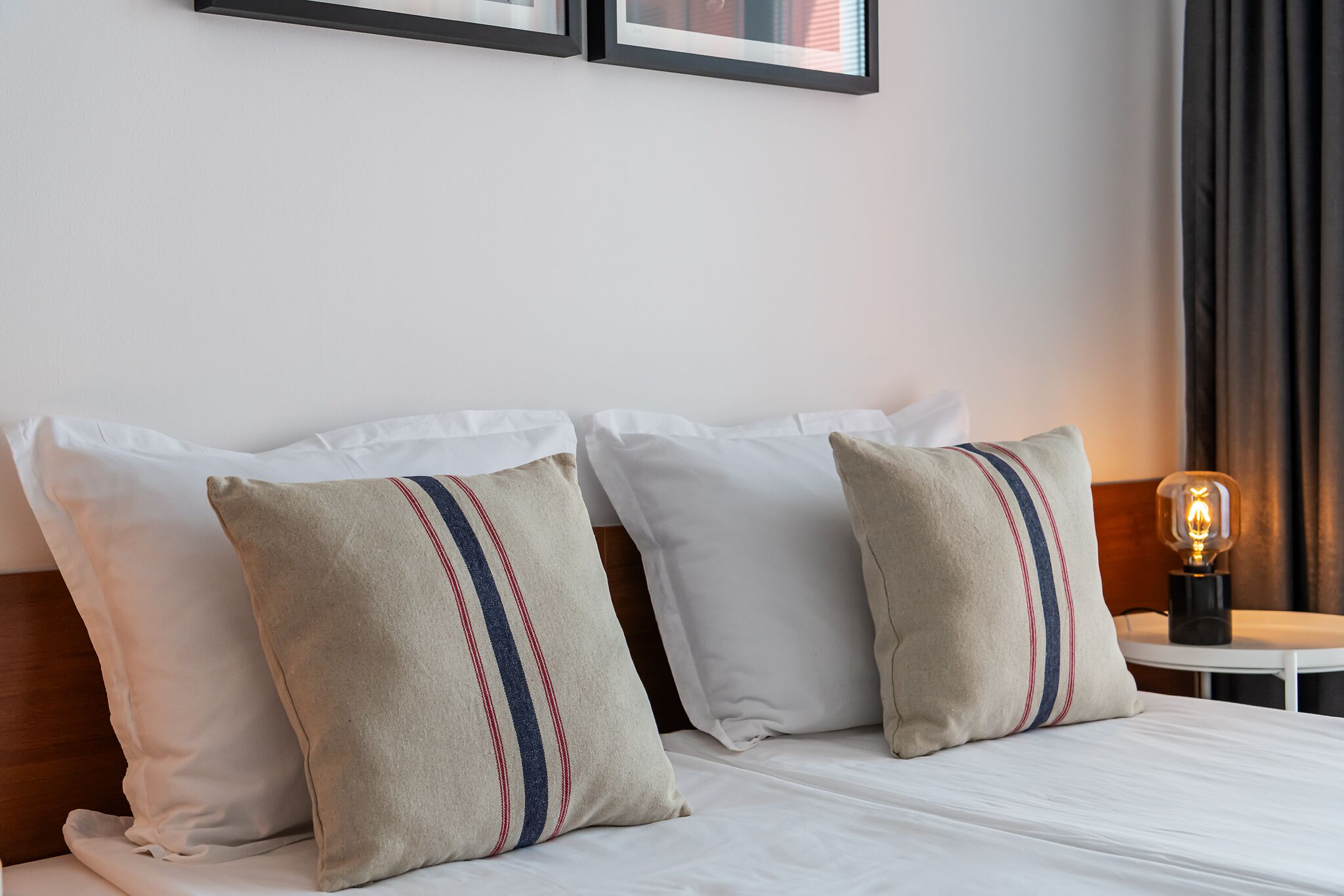 A neatly made bed with white linens and two pillows with red and blue stripes. There is a small table with a lamp beside the bed.