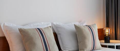 A neatly made bed with white linens and two pillows with red and blue stripes. There is a small table with a lamp beside the bed.