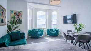 A spacious living room with teal sofas, a marble floor, and large windows offering a view of the sea. - St Catherine's View - 2 Bedroom - Panorama - Tenby (Tenby)
