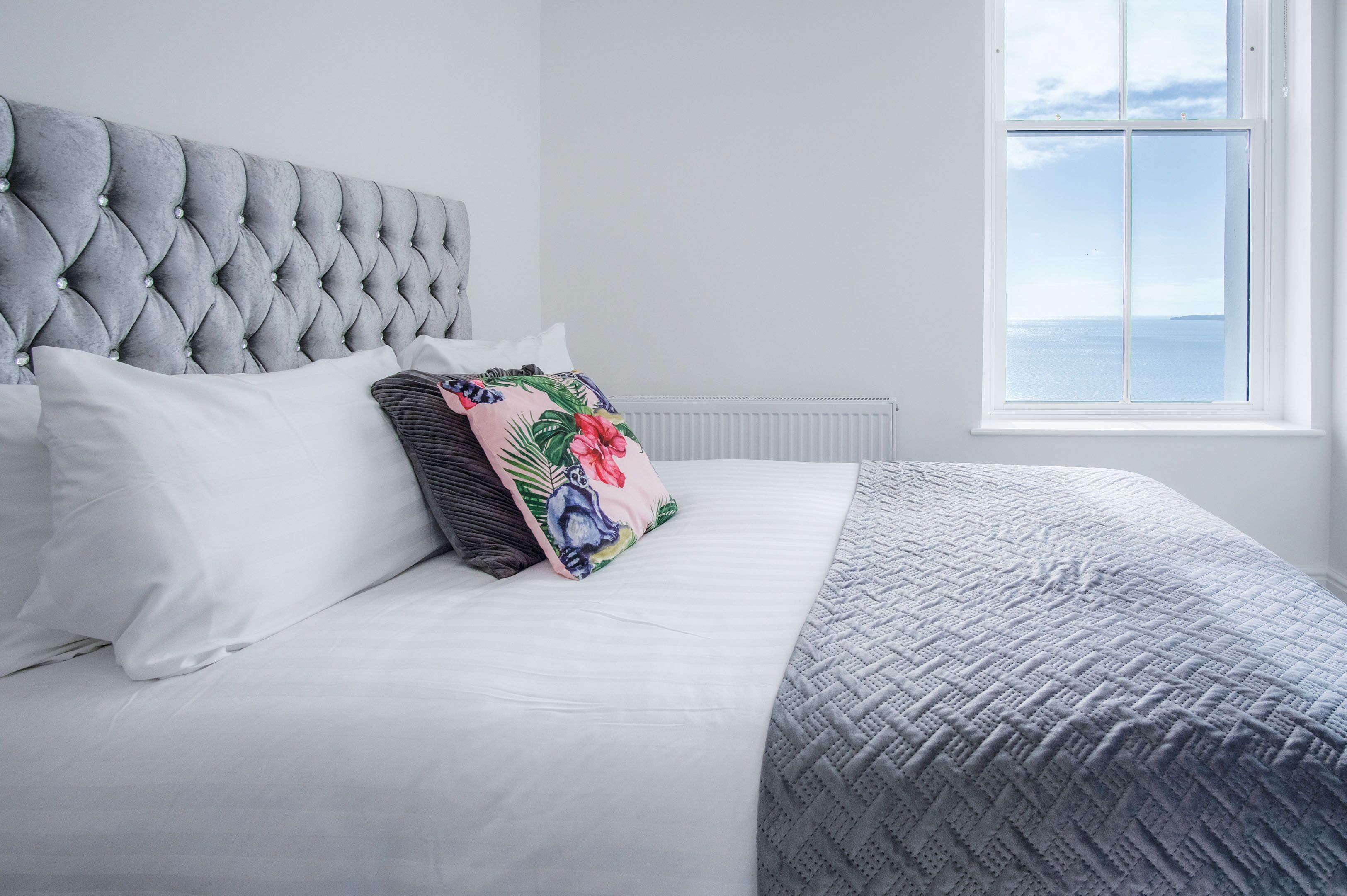 Una cama bien hecha con un cabecero gris, almohadas blancas y una almohada estampada con motivos florales.
