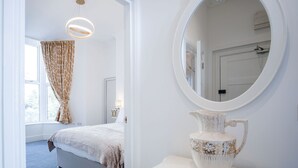 A bedroom with a bed, a bedside table with a vase, a window with curtains, and a mirror on the wall. - St Catherine's View - 2 Bedroom - Panorama - Tenby (Tenby)