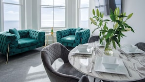 A modern living room with teal sofas, a marble table set for dining, and a large window with a view of the sea. - St Catherine's View - 2 Bedroom - Panorama - Tenby (Tenby)