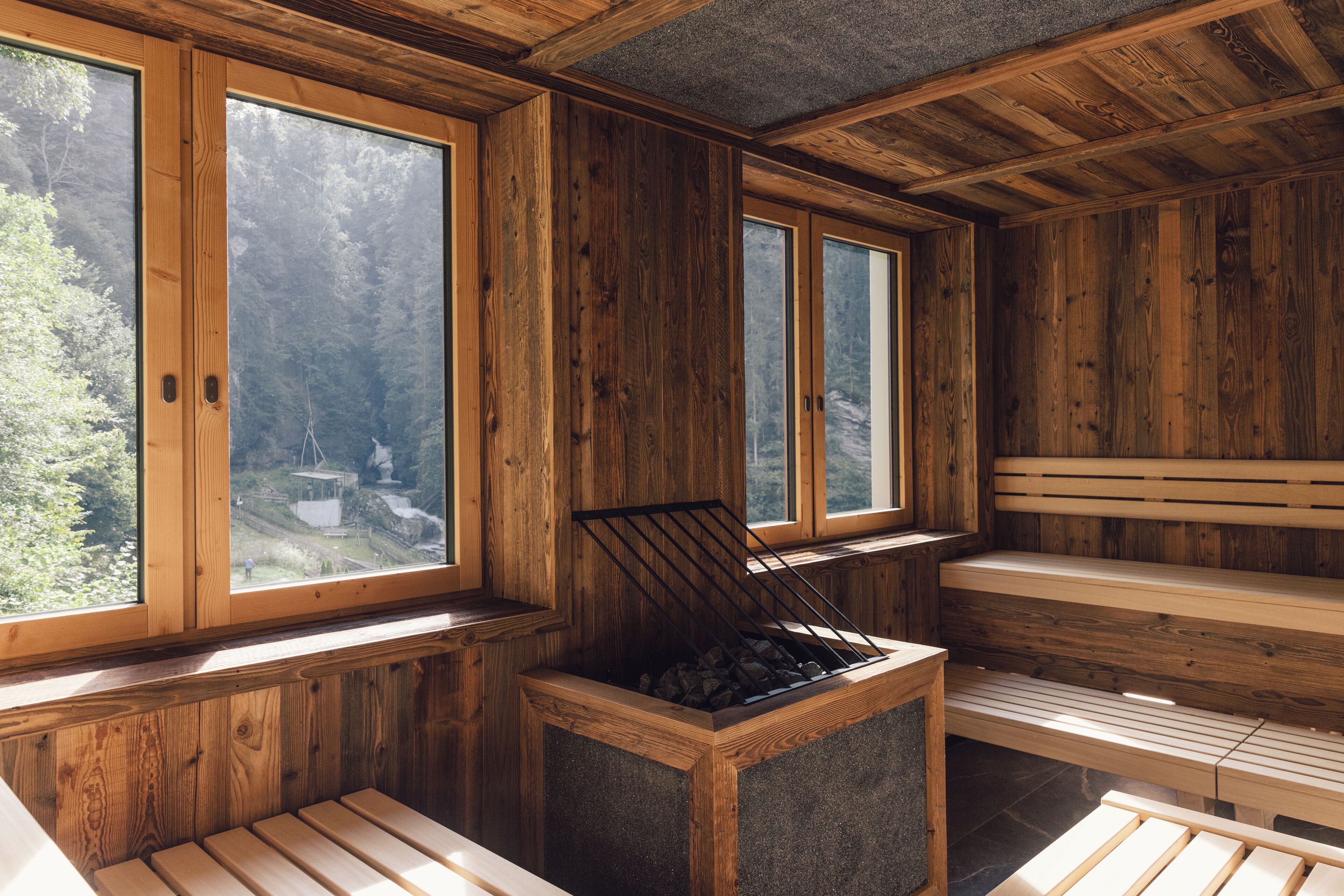 A wooden sauna with benches and a fire pit, large windows, and a view of a forest.