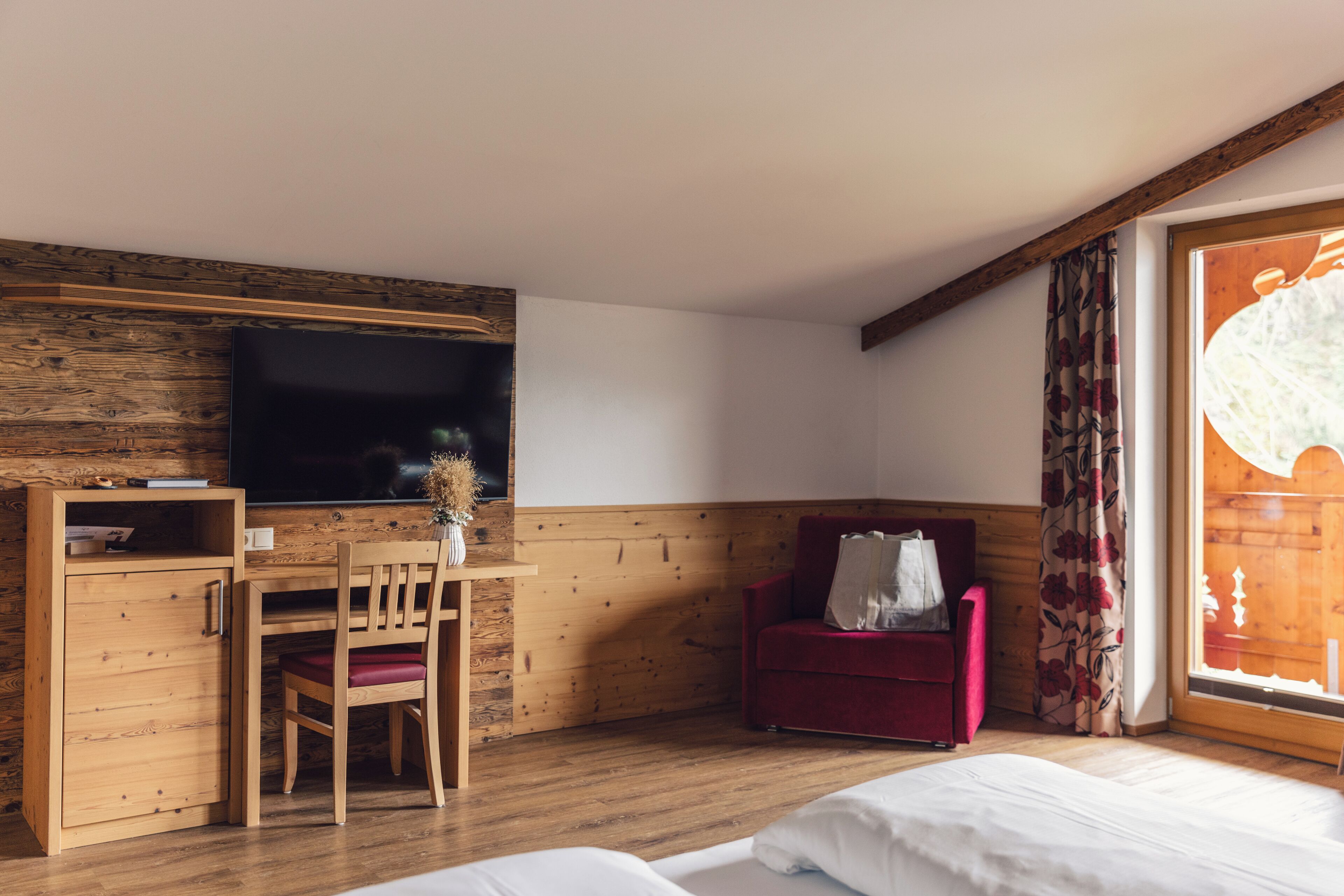 Een kamer met een houten bureau, een televisie, een rode stoel, een bed en een raam met gordijnen.