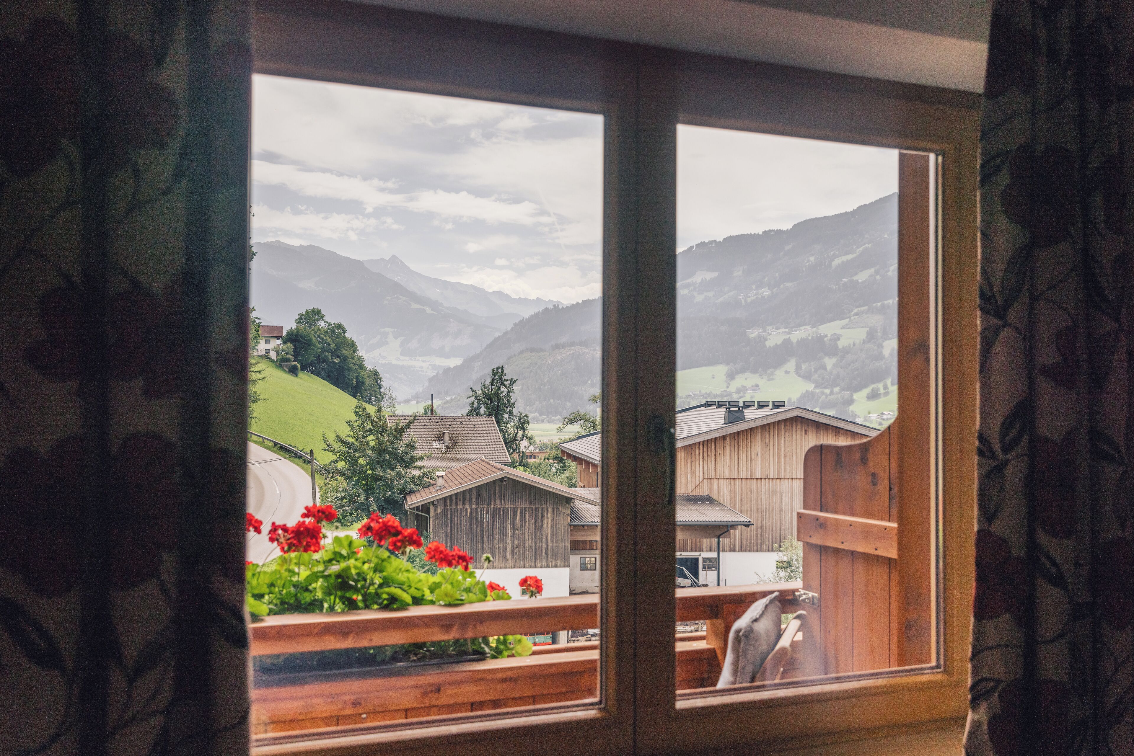 A view from a window with a wooden frame, overlooking a rural landscape with mountains, houses, and a wooden fence.