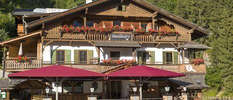 Un edificio tradicional de madera con balcones decorados con flores, base de piedra y mobiliario exterior con sombrillas.