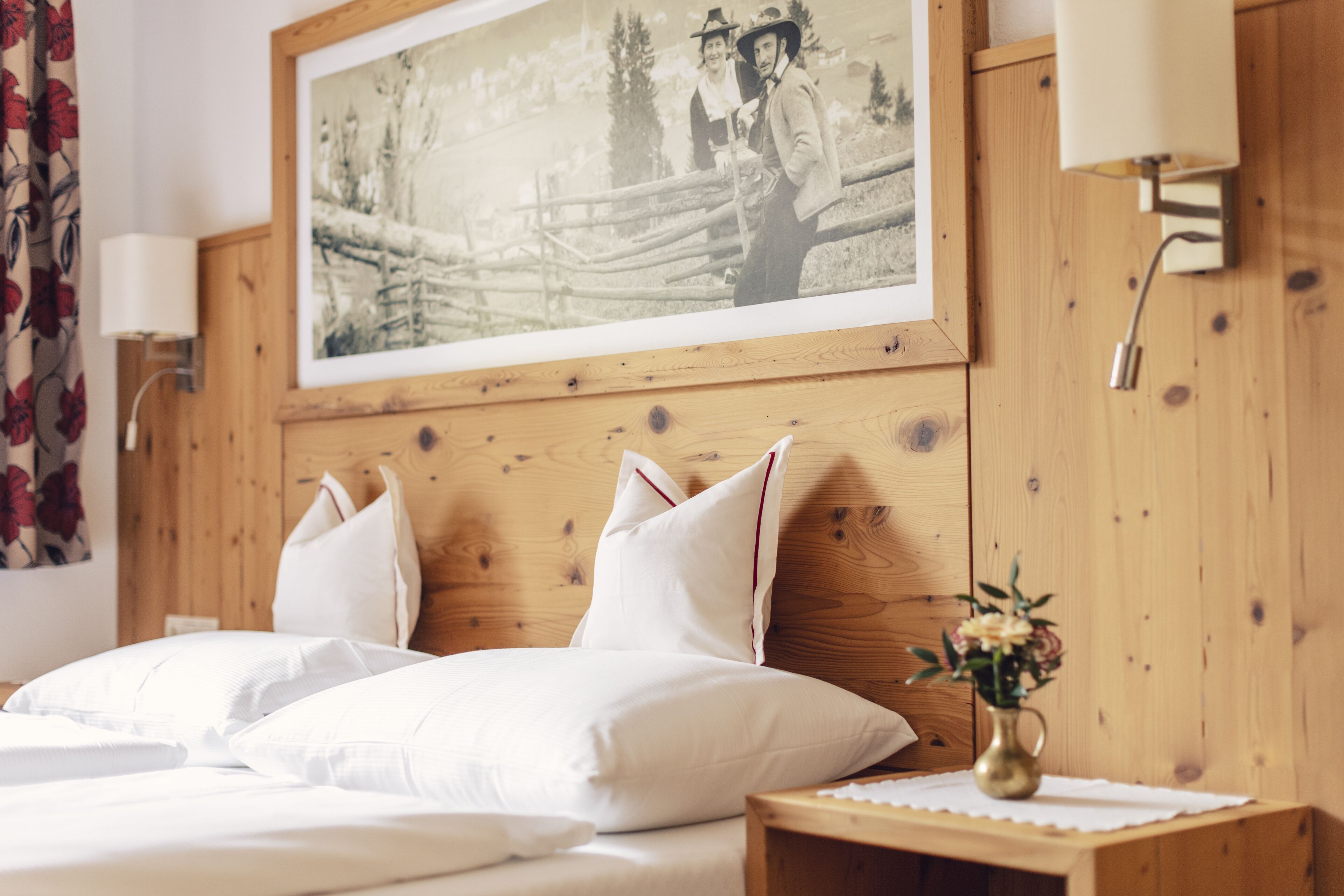 A neatly made bed with white linens and pillows, a framed photograph on the wall, and a small bedside table with a vase of flowers.