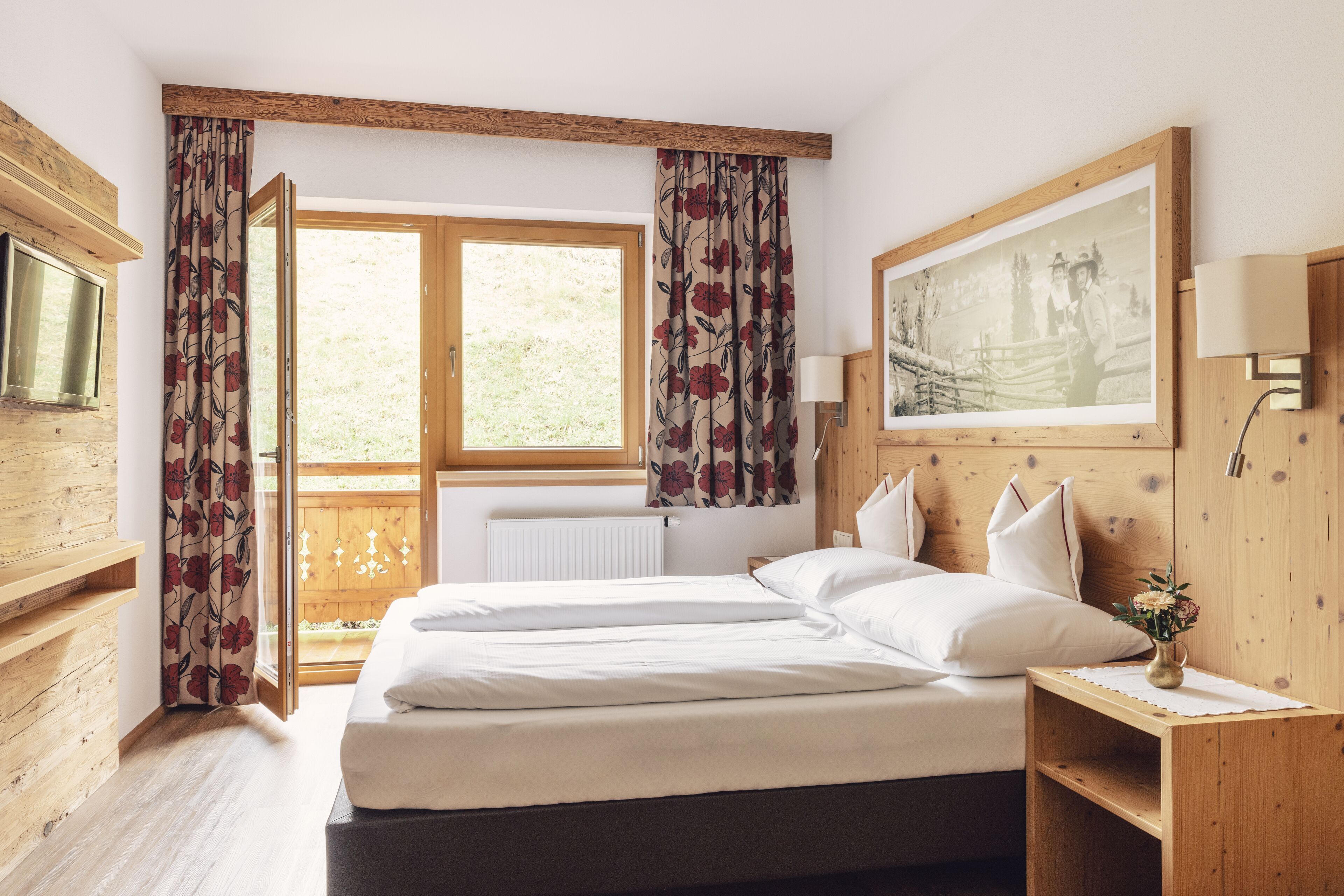 Een slaapkamer met een houten hoofdeinde, een groot bed met witte lakens, een nachtkastje van hout met een lamp en een raam met gordijnen.