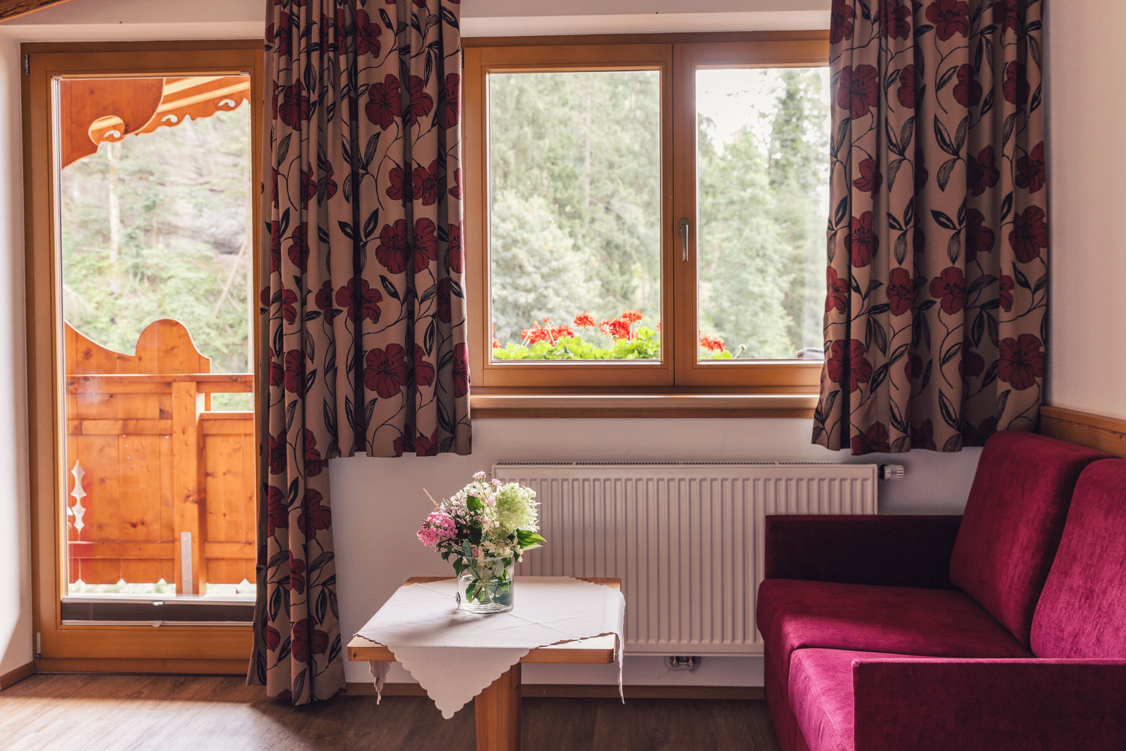 A cozy room with a wooden floor, a red sofa, a small table with a vase of flowers, and a window with curtains.