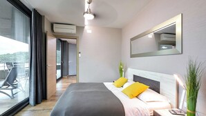 Habitación de hotel moderna con una cama grande, una mesita de noche con lámpara, un espejo y vistas al exterior.