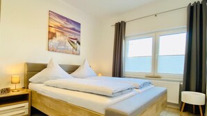 A modern bedroom with a wooden bed, a nightstand with a lamp, a small stool, and a window with curtains.