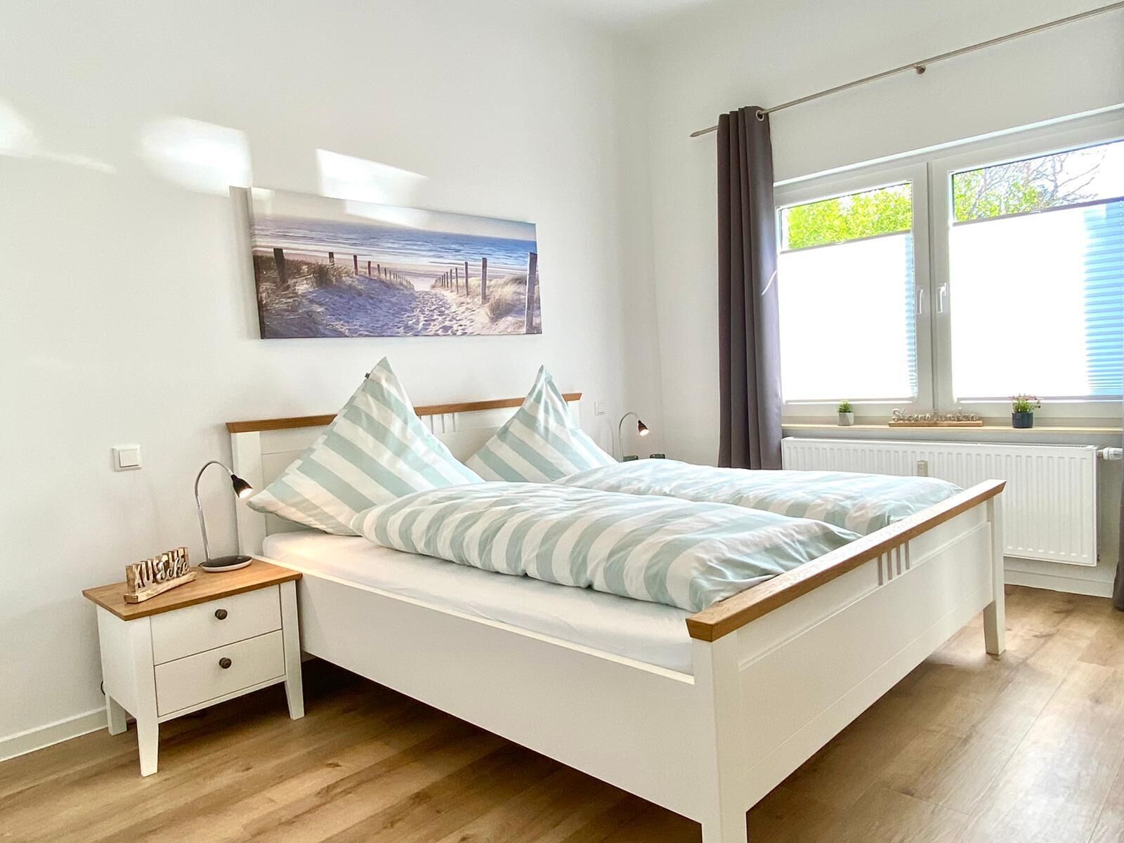 A bedroom with a bed, a nightstand, a lamp, a window with curtains, and a wall-mounted picture.