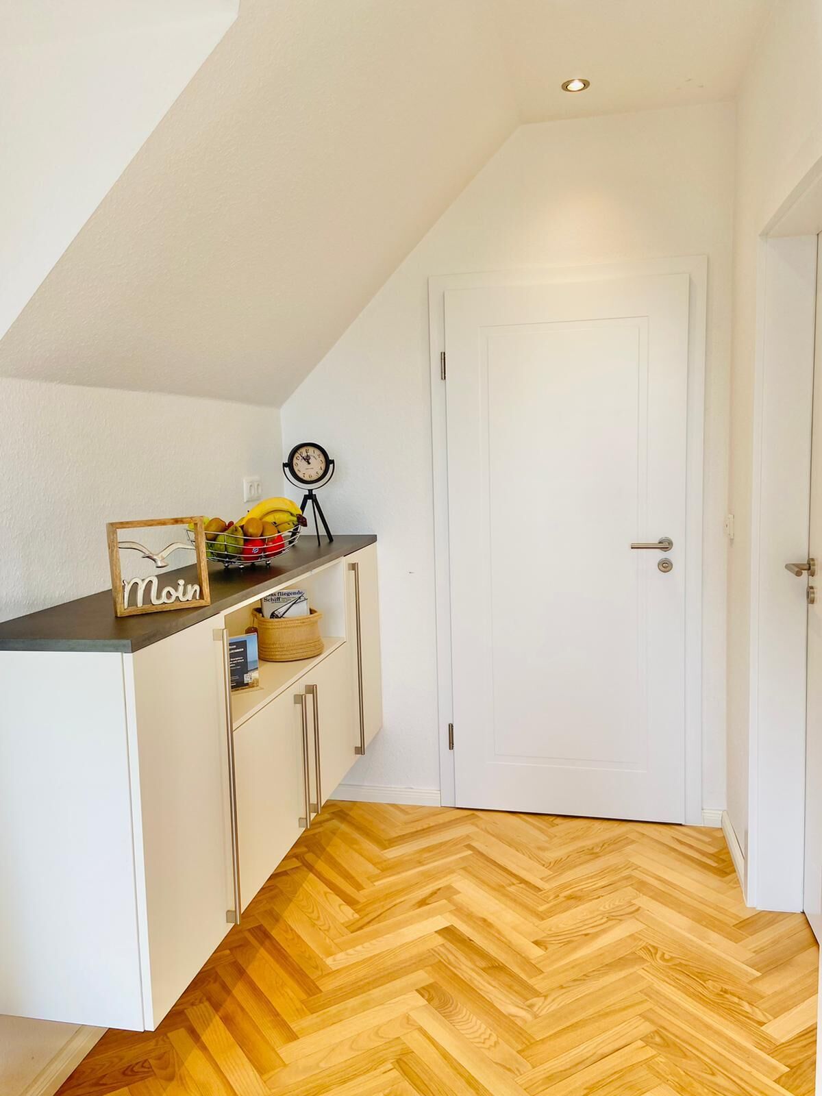 Une petite pièce bien éclairée, dotée d’un parquet en bois disposé en motif à chevrons, d’une porte blanche et d’un coin cuisine avec un panier de fruits et une horloge.