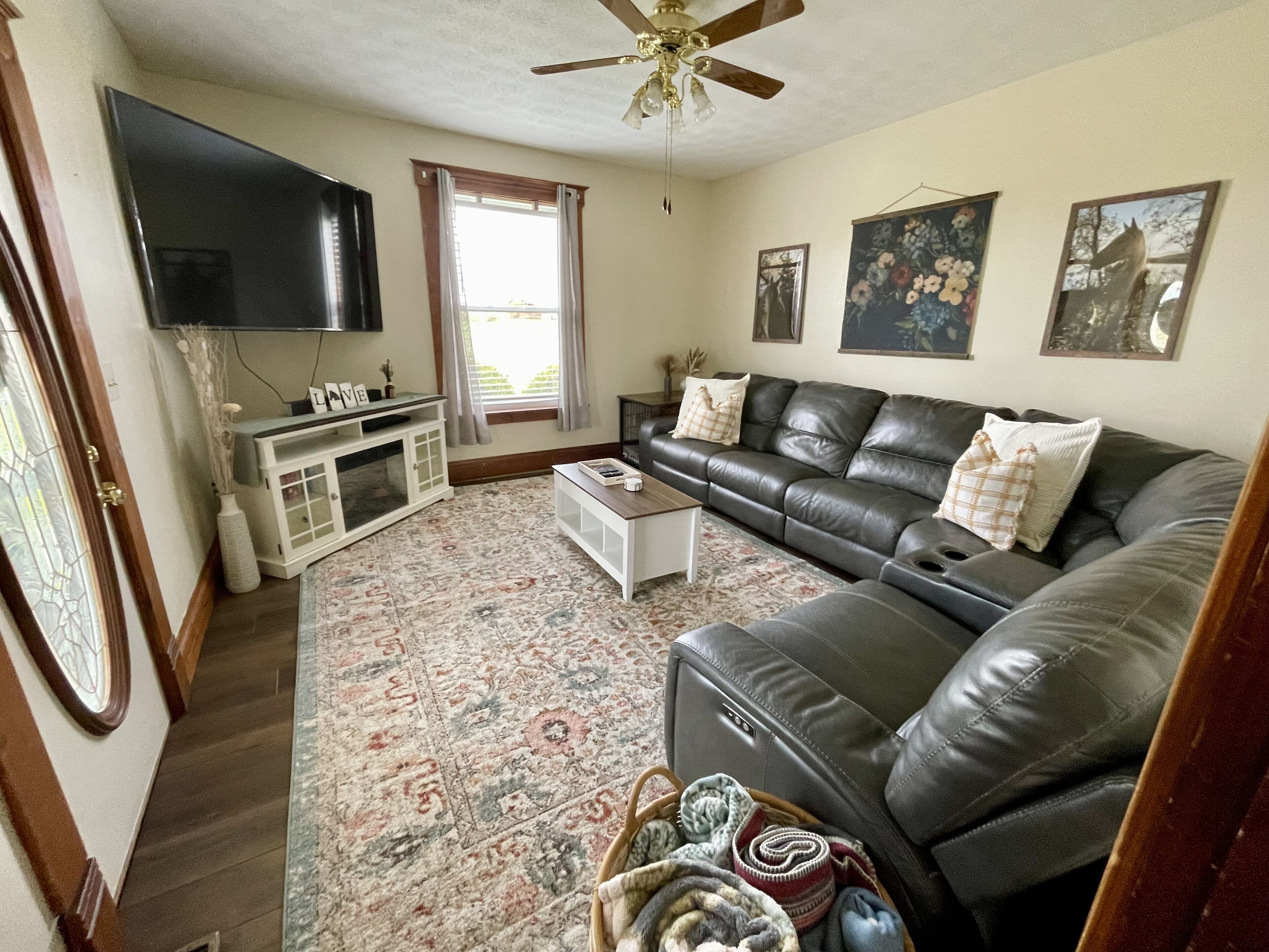 A living room with a leather sofa, a flat-screen TV, a ceiling fan, and a large window with curtains.