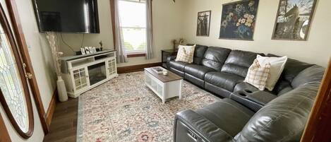 A living room with a leather sofa, a flat-screen TV, a ceiling fan, and a large window with curtains.