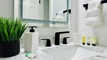 A bathroom with a white sink, black faucet, and a mirror above it. There is a potted plant, a towel rack with a towel, and a few toiletries on the counter.