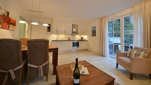 Une cuisine moderne avec des meubles blancs, un coin repas avec des chaises en osier, une table en bois avec une bouteille de vin, et un canapé près d’une grande fenêtre.