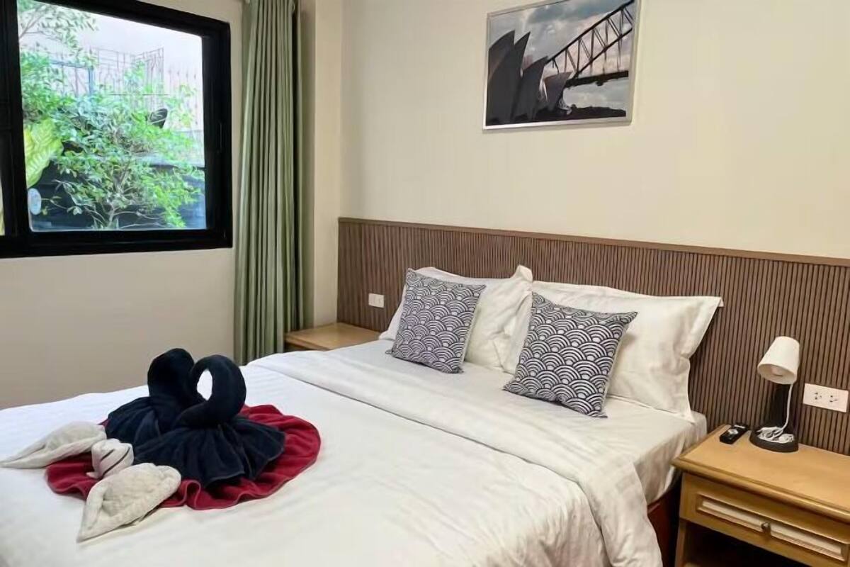 A hotel room with a bed, pillows, a nightstand, a lamp, a window with a view of greenery, and a painting on the wall.