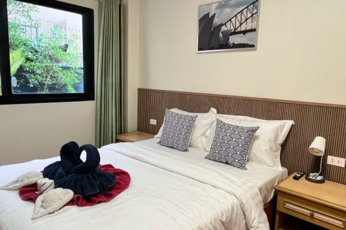 Une chambre d’hôtel avec un lit, des oreillers, une table de chevet, une lampe, une fenêtre donnant sur un paysage verdoyant et un tableau accroché au mur.