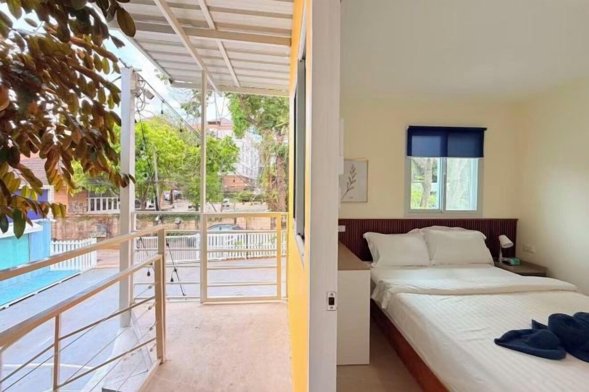 Habitación de hotel con cama, mesita de noche, ventana con persiana azul y vista a la calle y edificios.
