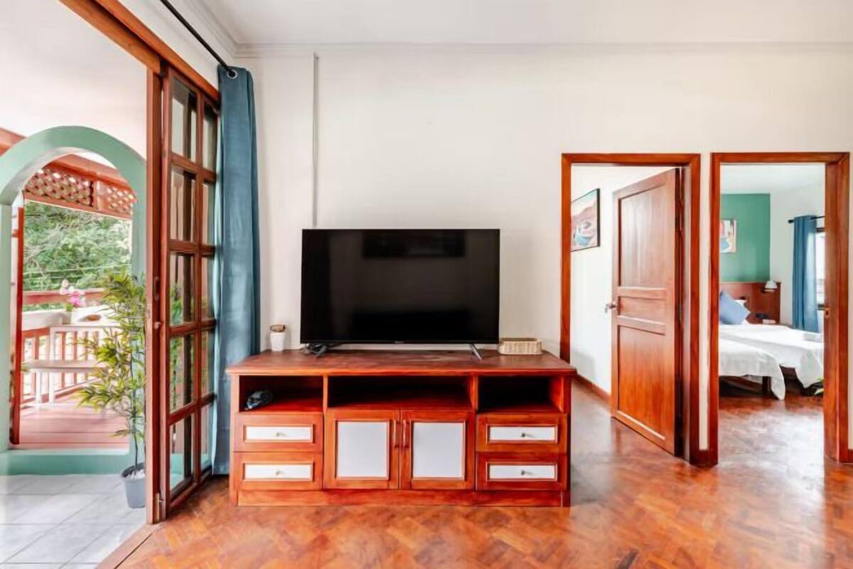 A room with a TV on a wooden stand, a bed with a blue headboard, and a door leading to another room.
