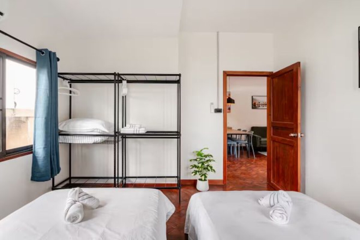 Une chambre d’hôtel avec deux lits, un lit superposé, une plante en pot et une porte en bois donnant sur un coin repas.