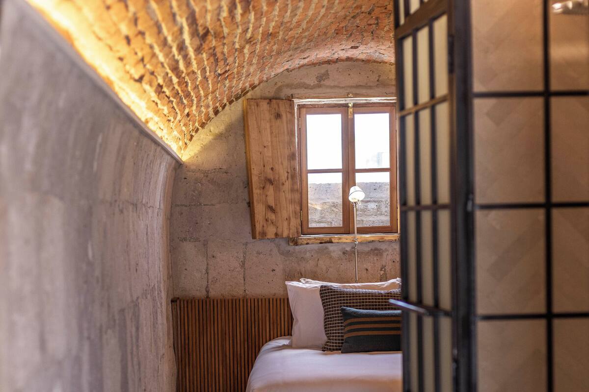 A narrow, rustic room with a sloped ceiling, a wooden bed, a window, and a tiled floor.