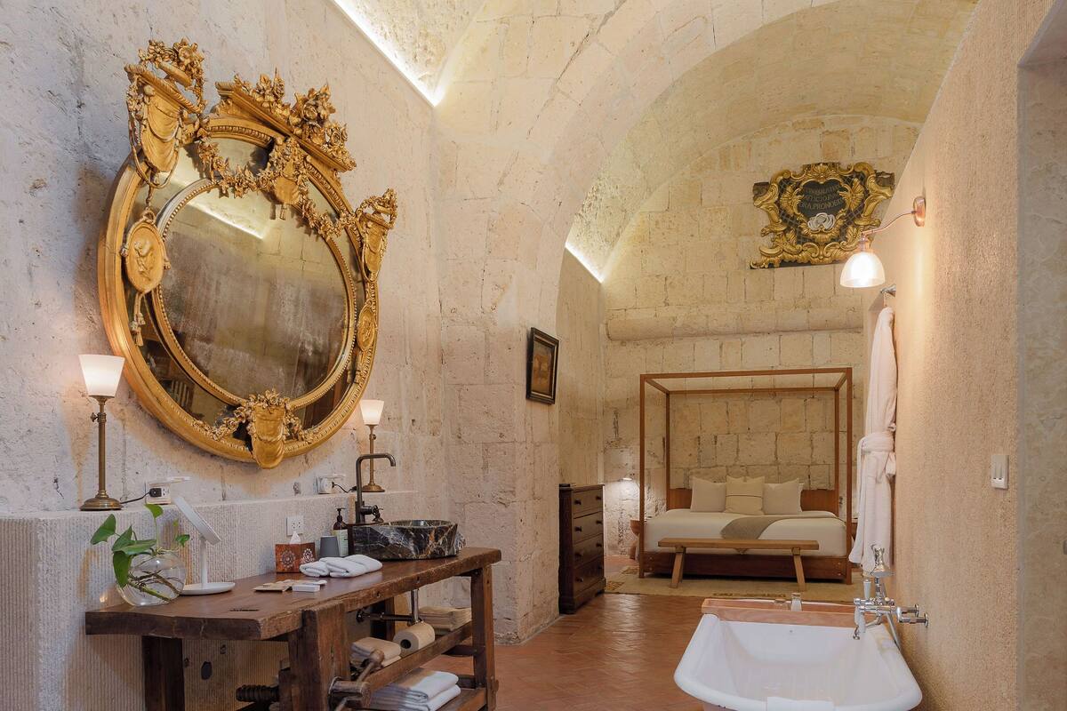 A bathroom with a bathtub, a large ornate mirror, a wooden vanity, and a bed with a wooden headboard.