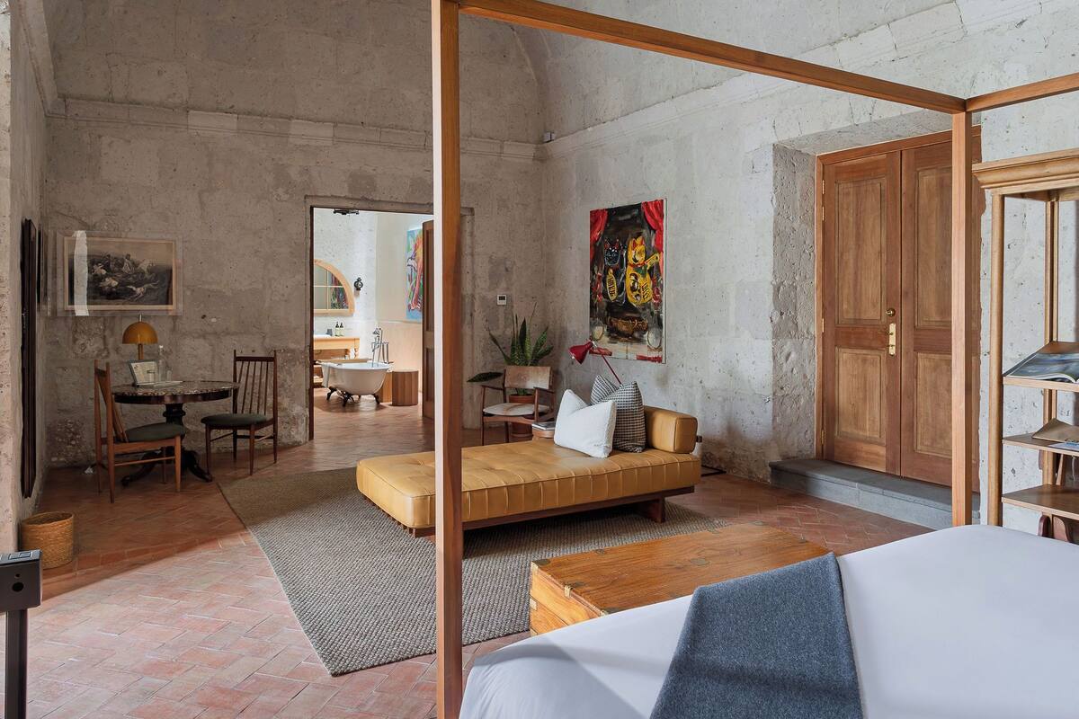 A room with a bed, a sofa, a wooden cabinet, a chair, a table, a painting, and a bathroom visible through an open door.