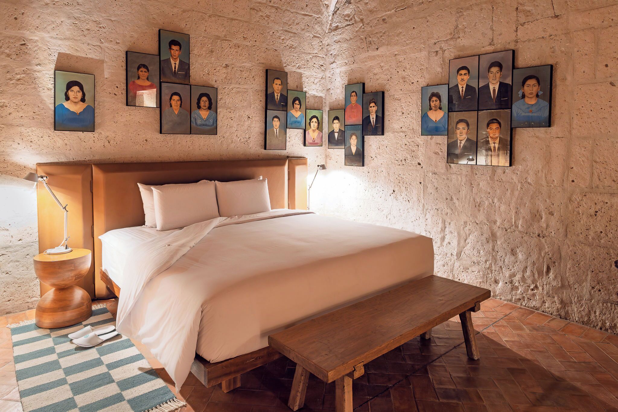 A bedroom with a bed, bedside table, lamp, bench, and a wall with portraits.