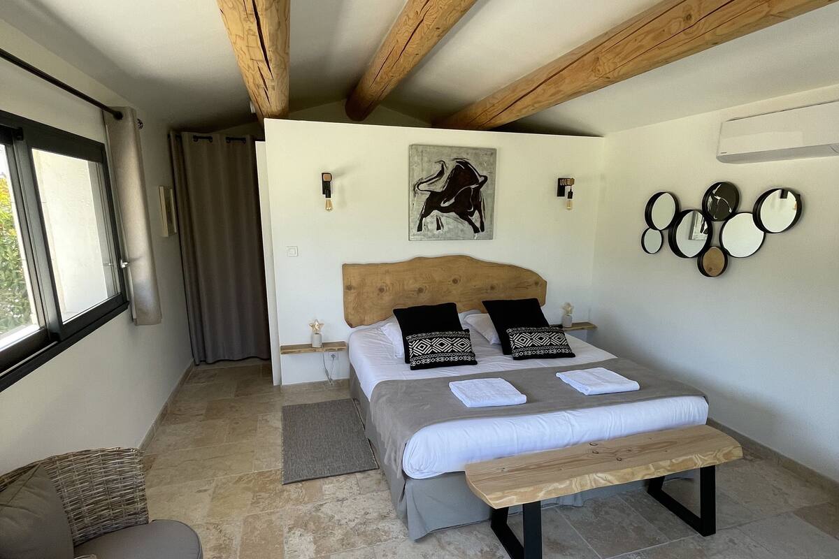 A bedroom with a bed, a bench, a wicker chair, a wall-mounted mirror, and a ceiling fan.