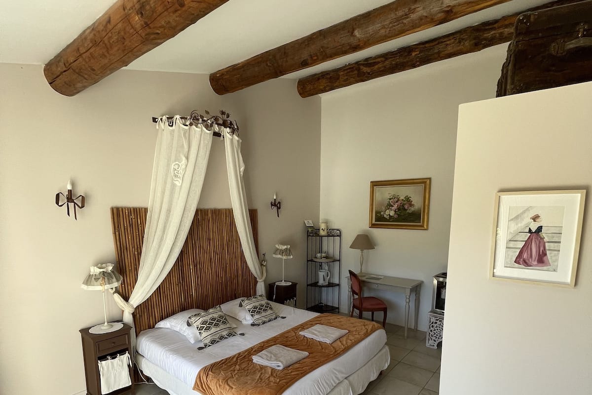 A bedroom with a large bed, wooden beams, a nightstand, a desk, and a chair.