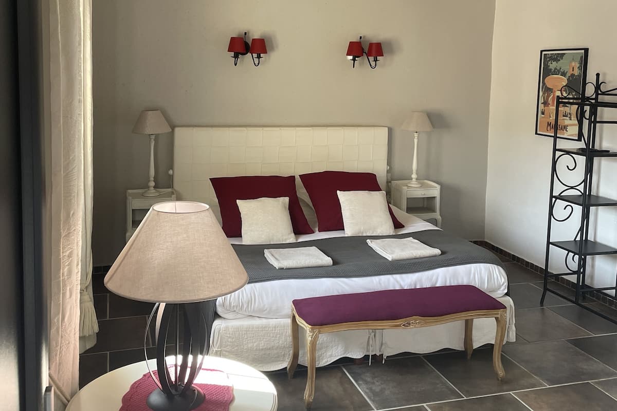 A bedroom with a bed, bedside lamps, a small table with a lamp, a bench, and a wall-mounted shelf.