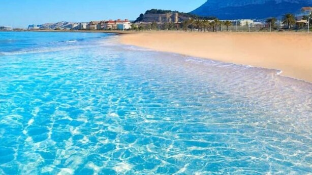 A beach with clear blue water, sandy shore, and buildings in the background. - Casa Solanilla Entre mer et Golf (La Sella)
