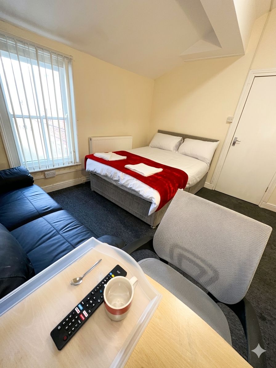 Une chambre d’hôtel comprenant un lit, un canapé, une table avec une télécommande et une tasse, ainsi qu’une fenêtre avec des stores.