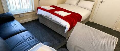 Une chambre d’hôtel comprenant un lit, un canapé, une table avec une télécommande et une tasse, ainsi qu’une fenêtre avec des stores.