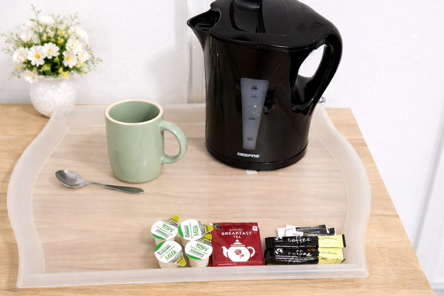 A coffee maker, a mug, and a tray with milk cartons, a red bag, and coffee packets on a wooden table.