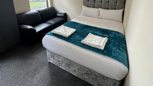 A bedroom with a bed, a black leather sofa, a window, and a view of buildings outside. - Macclesfield Lodge (Macclesfield)