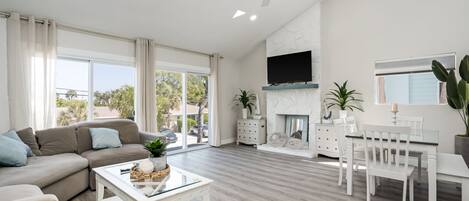 Amplia sala de estar con un ventanal grande, chimenea, ventilador de techo y comedor con mobiliario blanco.